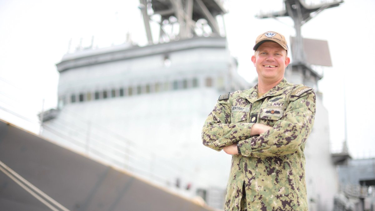 A soldier stands arms crossed in front of an aircraft carrier.
