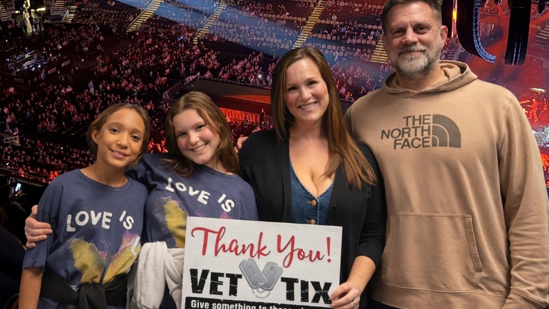 A family of four holds a Vet Tix thank you sign at a concert venue.