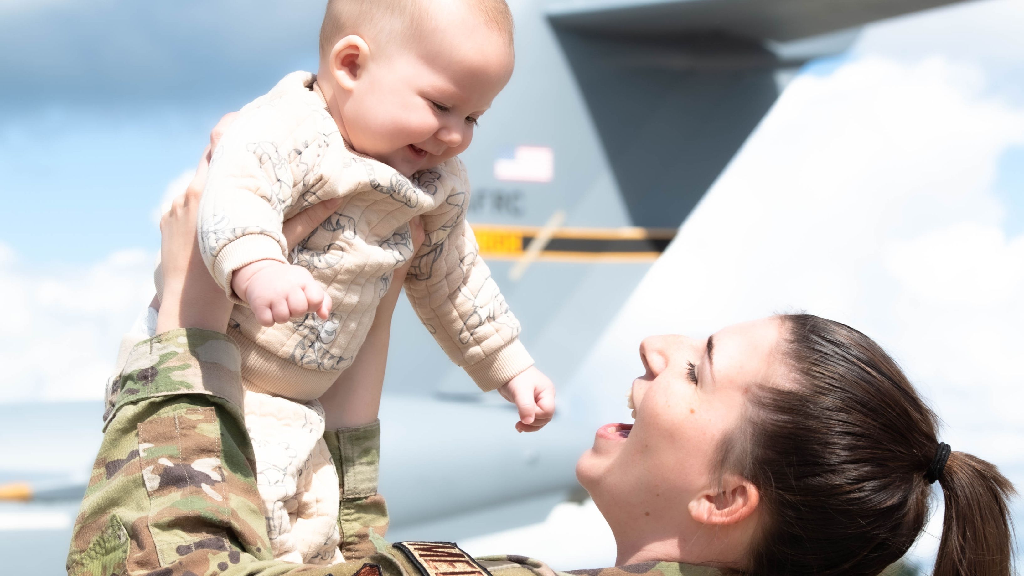 Uniformed mom holds up happy baby on the tarmac of an airfield.