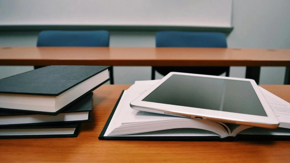 Tablet and open book on classroom desk with stack of closed books in background