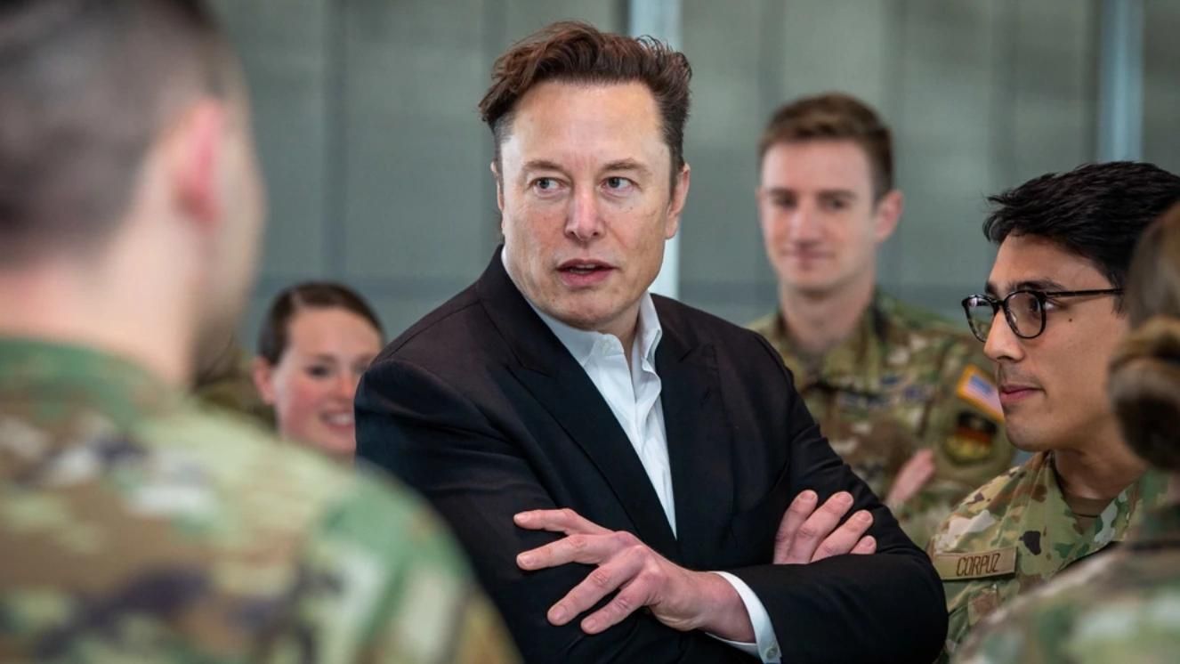 Elon Musk speaking with Air Force personnel in uniform indoors.