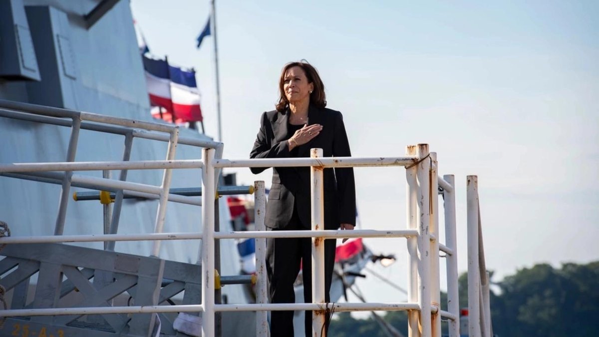 Kamala Harris on Navy ship deck with flags in background, hand over heart