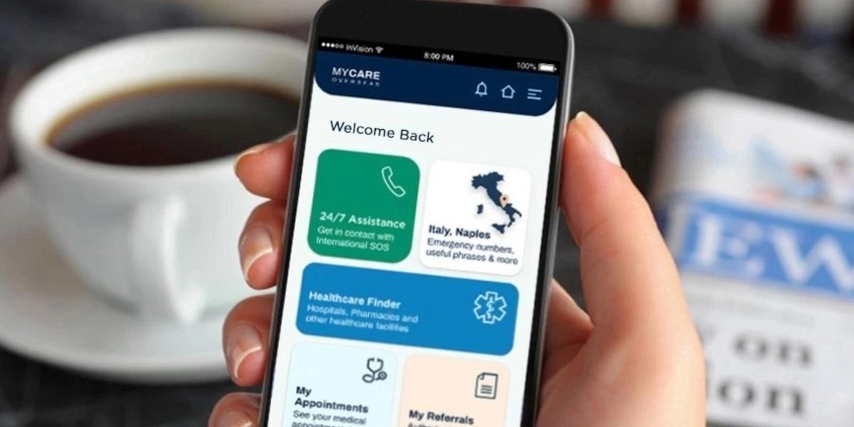 Person holding smartphone displaying TRICARE app with eligibility options, coffee cup and magazine in background.