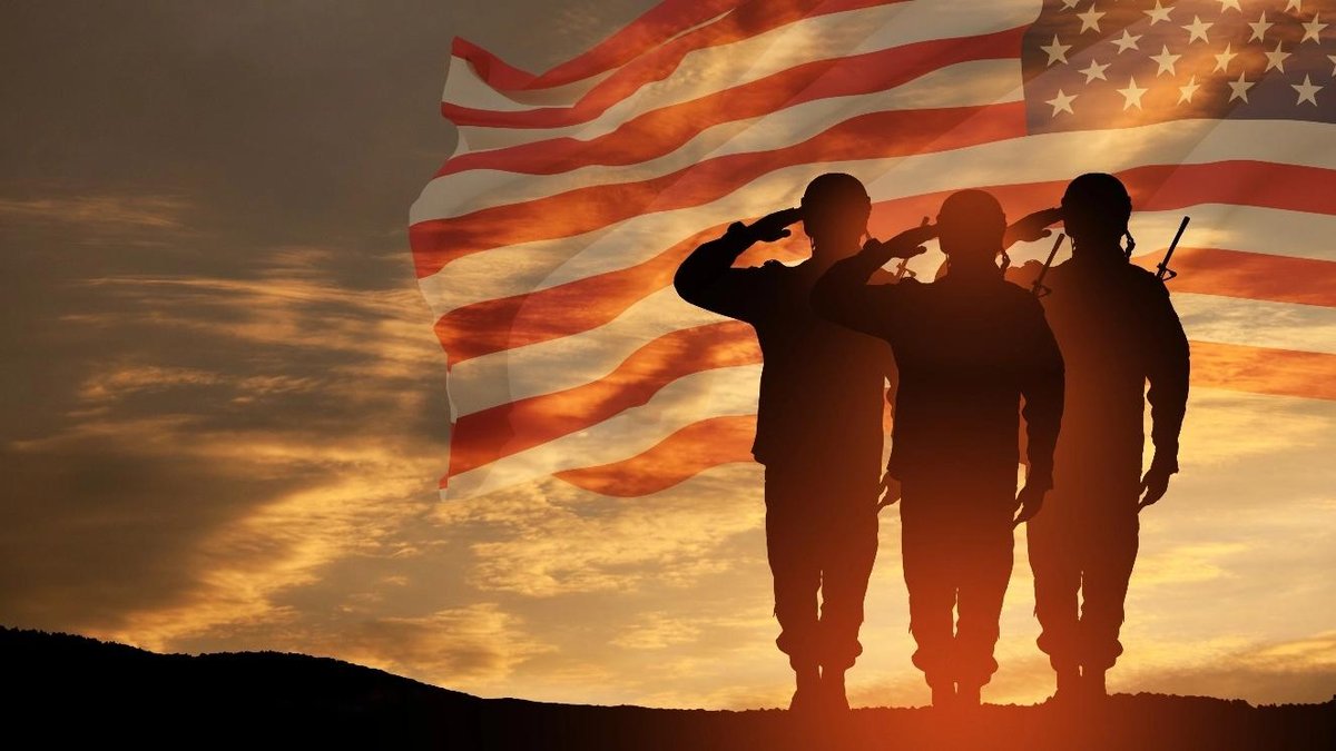 Silhouetted soldiers saluting with American flag against sunset sky.