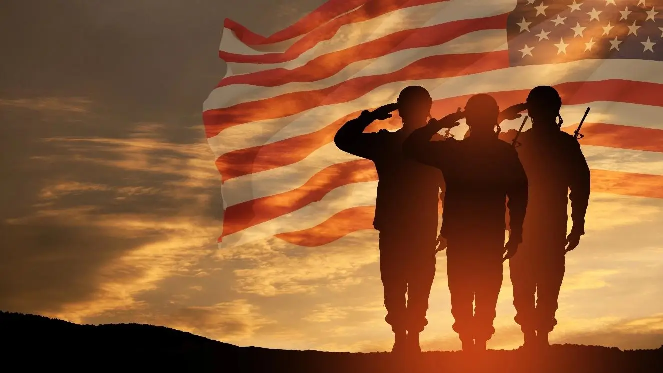 Silhouetted soldiers saluting with American flag against sunset sky.