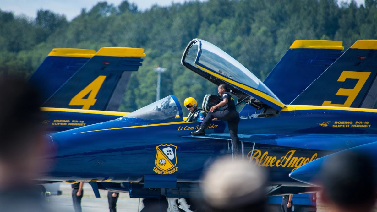 A Blue Angel Serviceperson stands on a wing of a fighter jet talking to the pilot.