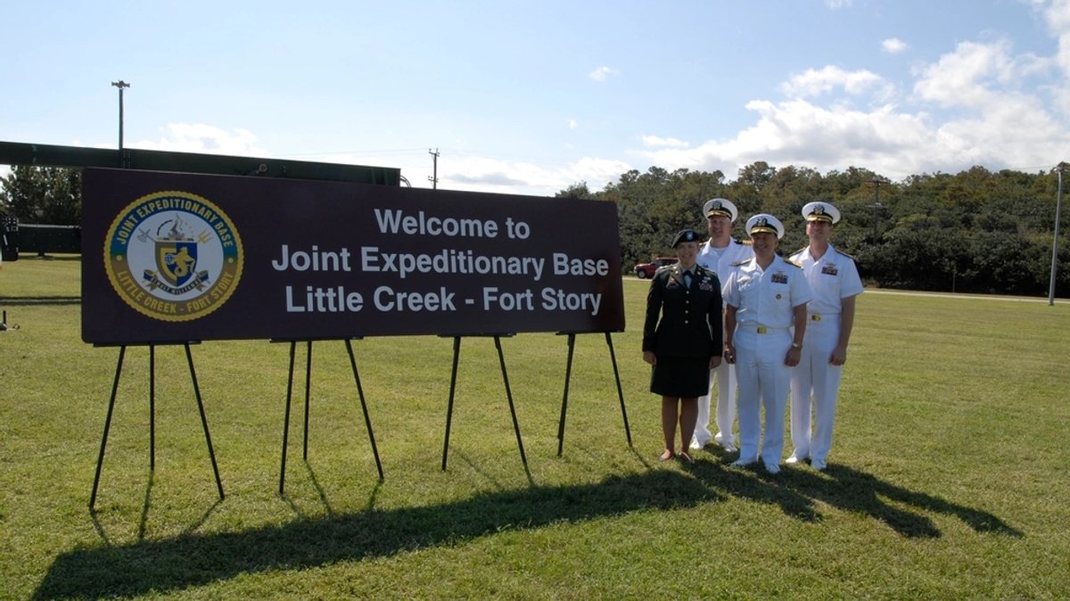 Formally dress soldiers stand next to a Welcome sign for Little Creek-Fort Story.