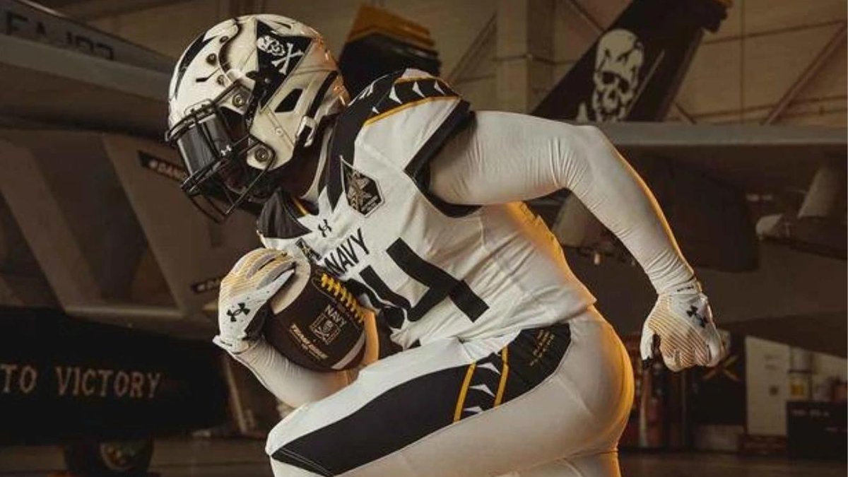 Navy Midshipman in Jolly Rogers-themed uniform posing with football in aircraft hangar.