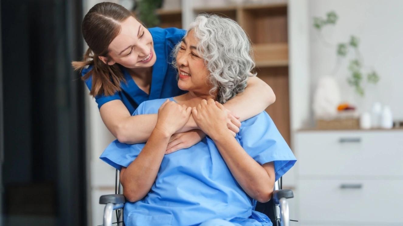 Caregiver in blue scrubs hugging elderly woman in wheelchair indoors