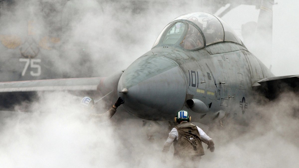 Two Air Force soldiers guide an F-14 through smoke and steam.
