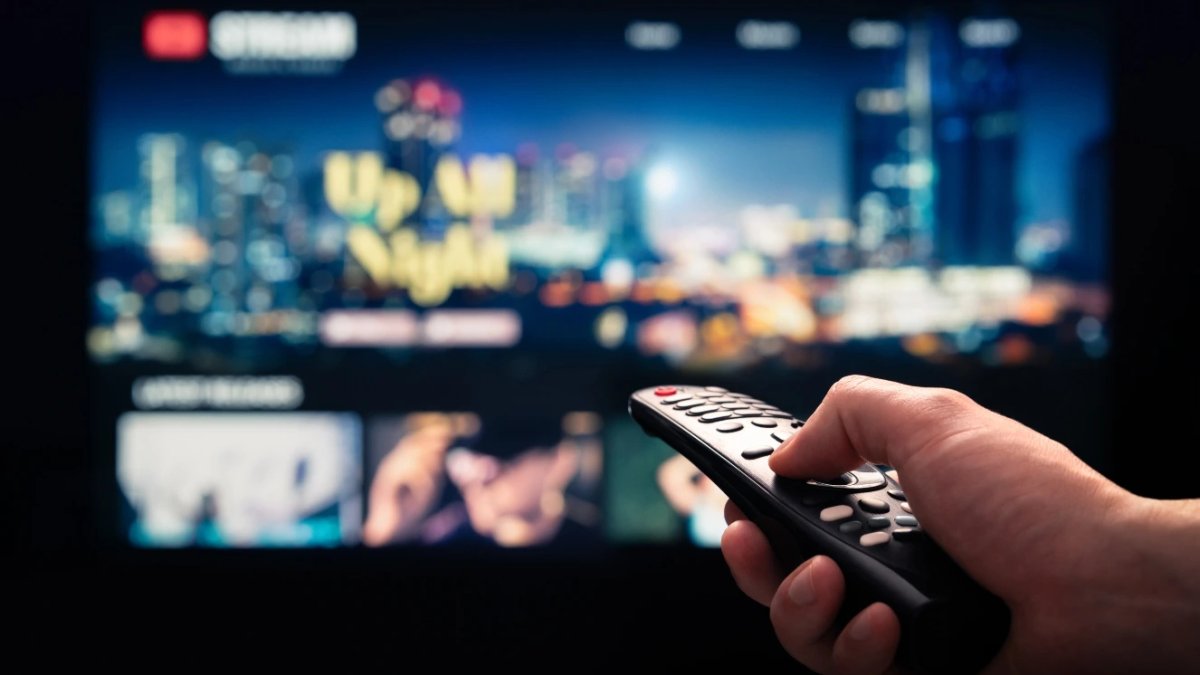 Hand holding remote control in front of TV displaying streaming service interface.