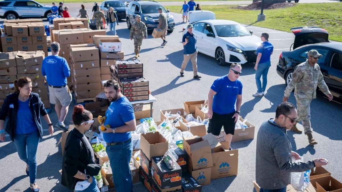 Volunteers and service members distribute food and supplies at military charity event in parking lot.