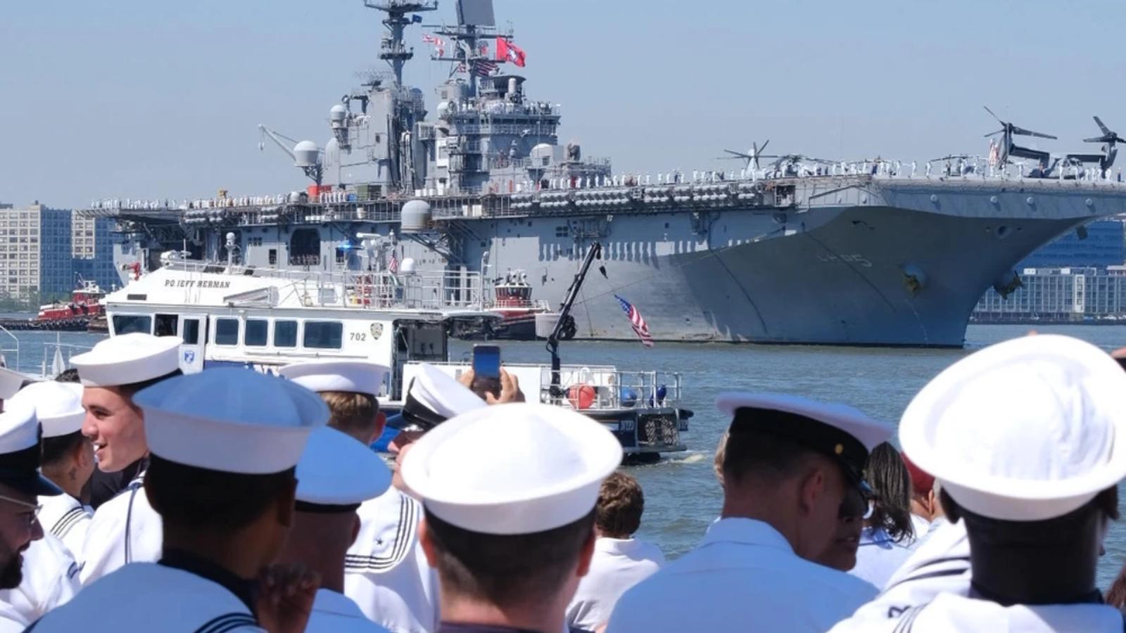 Navy sailors gathered near Arleigh Burke destroyer with aircraft carrier in background.