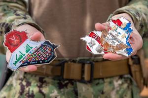 Two hands holding two oddly shaped challenge coins. They are in the shape of the countries Iraq and Afghanistan.
