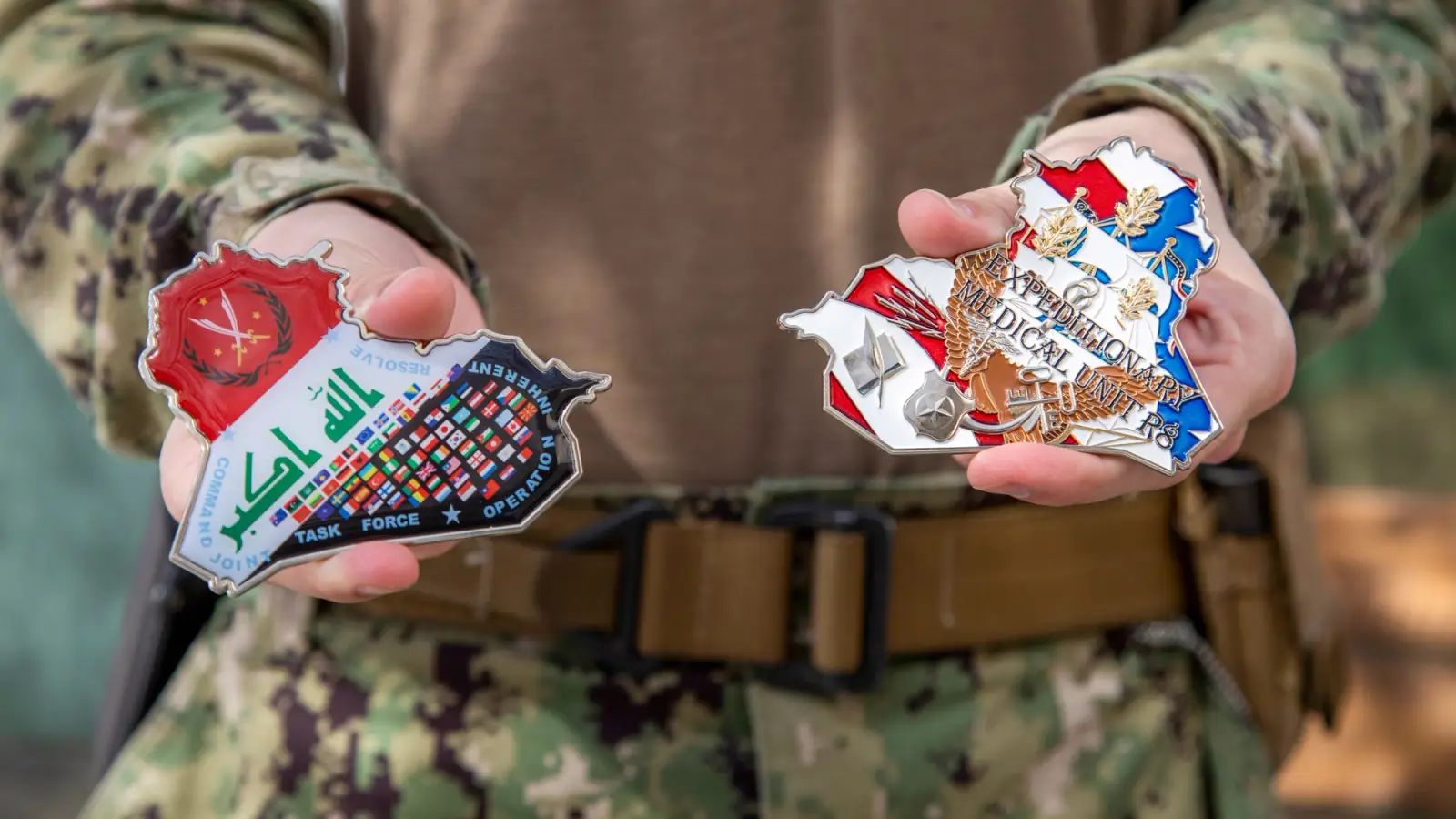 Two hands holding two oddly shaped challenge coins. They are in the shape of the countries Iraq and Afghanistan.