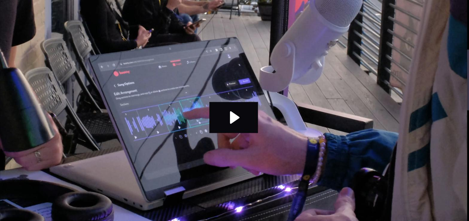 Photo of a laptop screen showing the Boomy song editor with an 'Edit Arrangement' waveform, overlaid with a video play button, while a hand points at the screen near a microphone at what appears to be a live event.