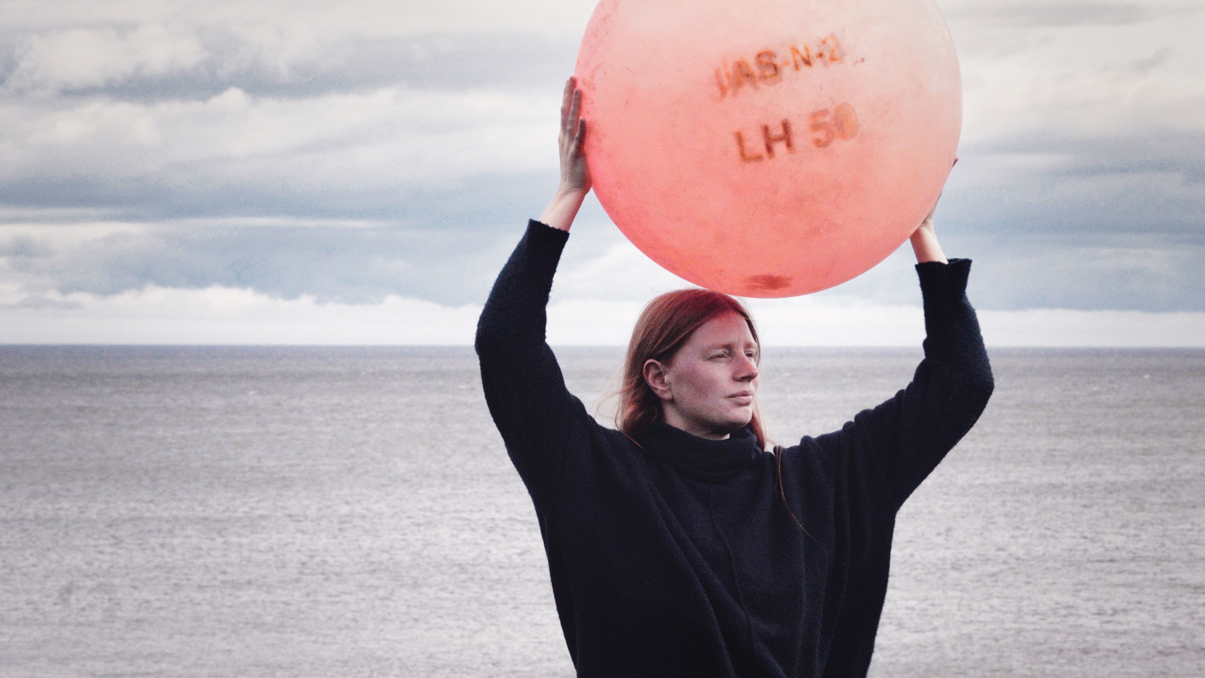 A woman in black lifts a red buoy above her head with the sea behind.