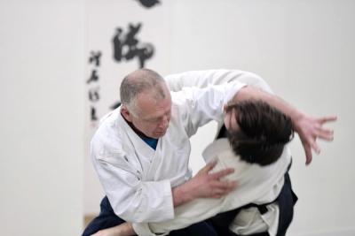 Aikido instructor performing iriminage throw on a student with calligraphy scrolls visible