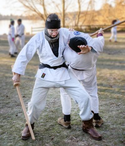 Dynamic aikido throw with weapons during outdoor training session with practitioners in the background