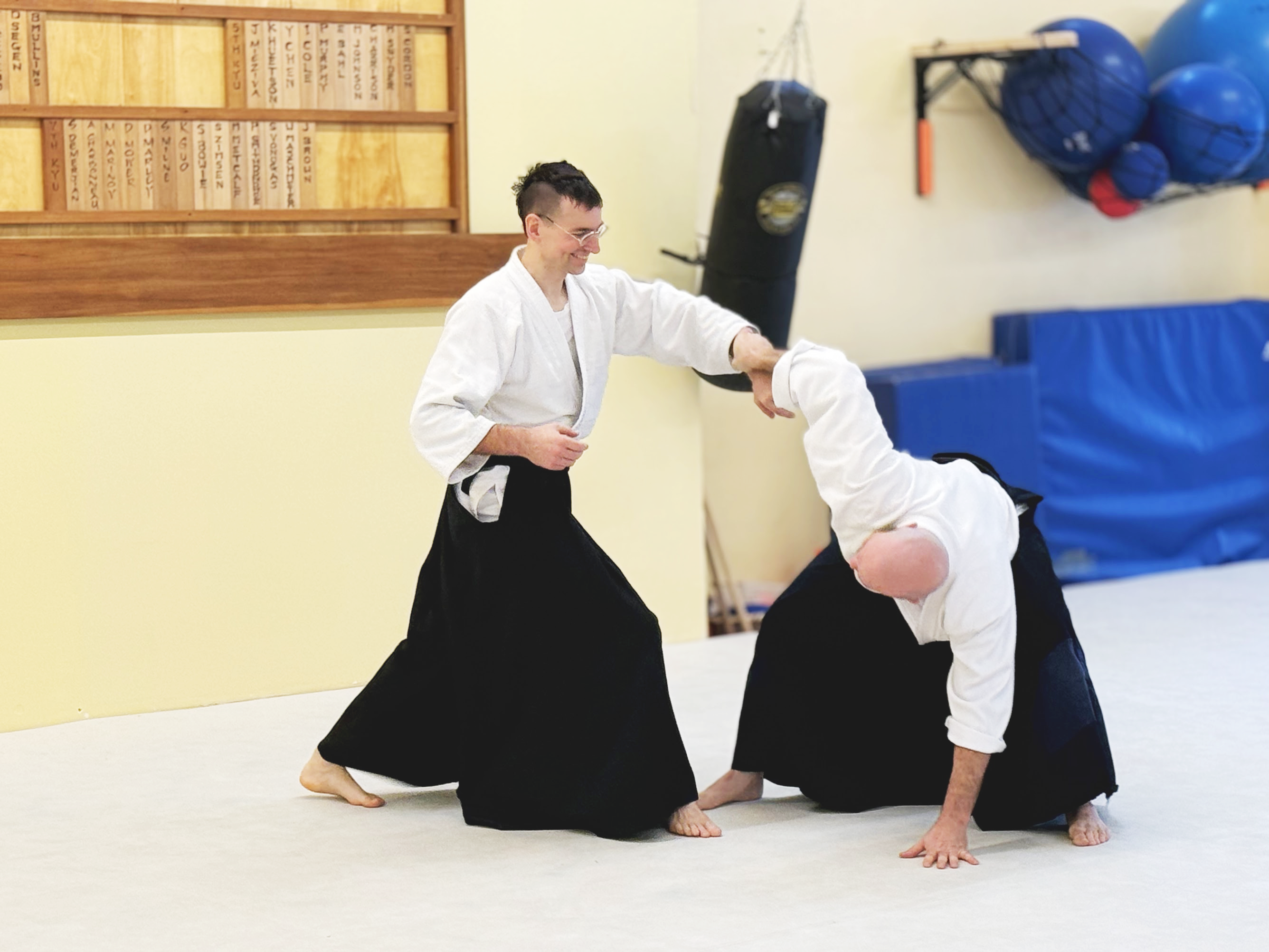 Two aikido students in white gi and black hakama practicing a throw on the dojo mat.