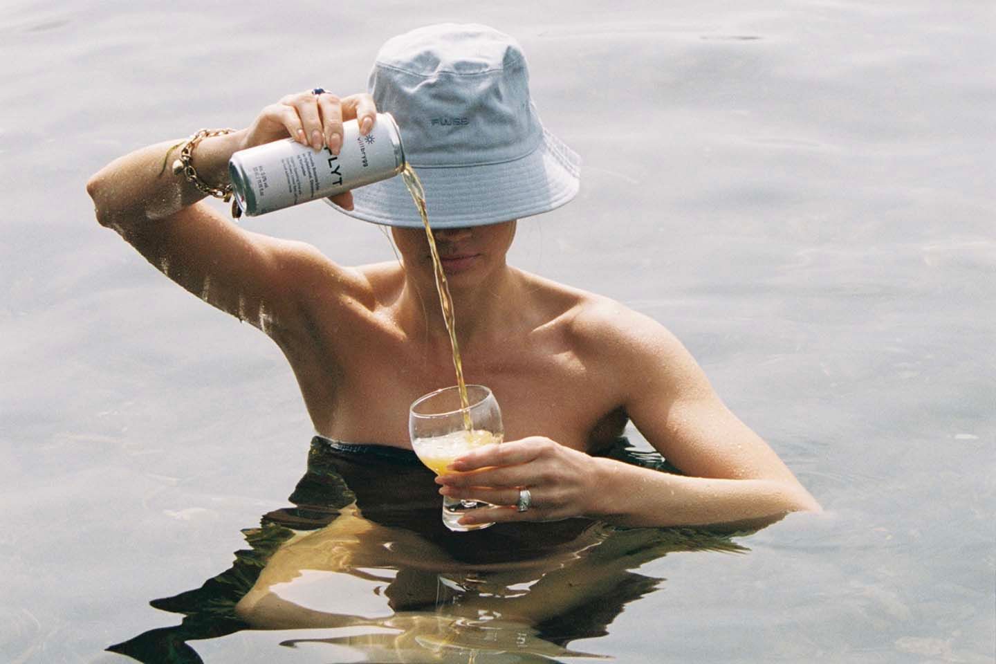 På bildet ser man en kvinne stå i vannet og helle opp drikke i et glass.