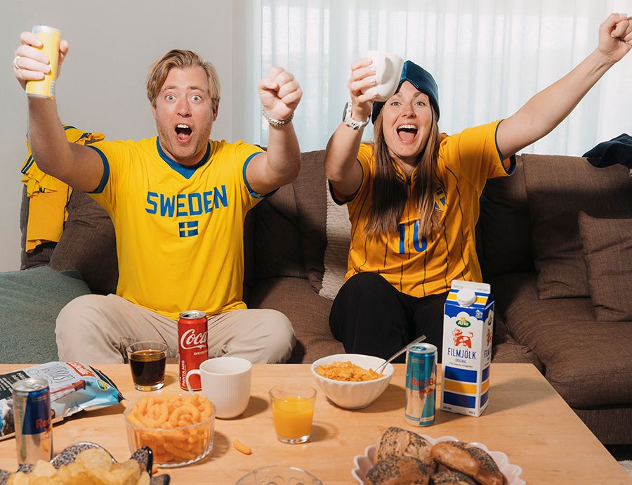 På bilden syns två personer i gul-blåa matchtröjor sitta i en soffa, de ser ut att ropa glatt och har snacks på bordet framför sig