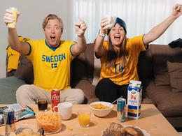 På bilden syns två personer i gul-blåa matchtröjor sitta i en soffa, de ser ut att ropa glatt och har snacks på bordet framför sig