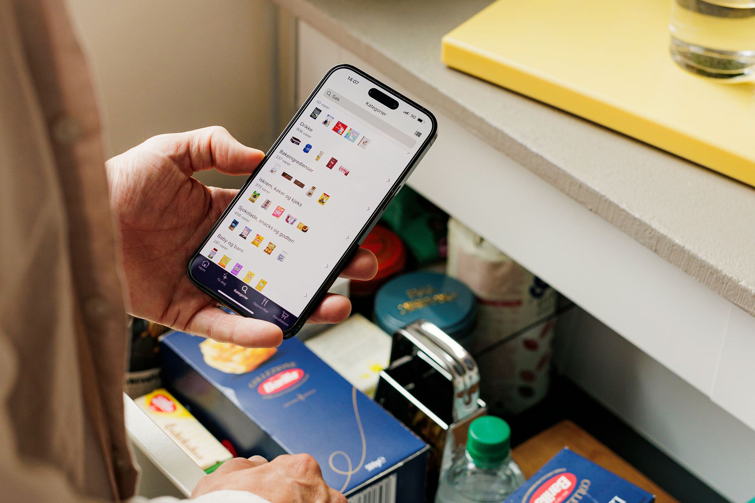 På bildet ser man en uttrekksbar kjøkkenskuff halvfull med tørrvarer. Over skuffen ser man også en hånd som holder en mobil, og bruker Odas app for å planlegge matinnkjøpene.