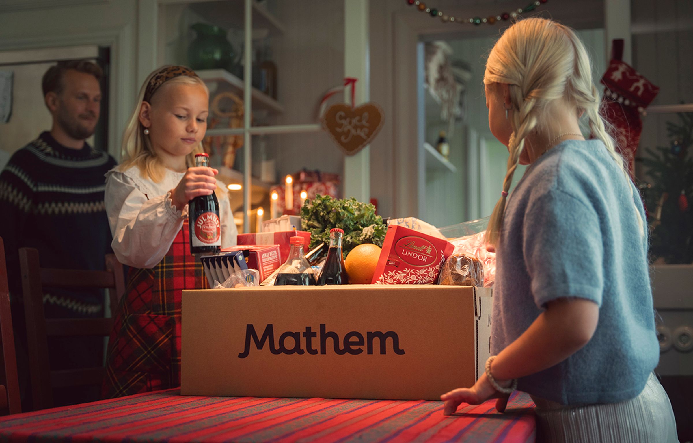 På bilden syns två barn packa upp varor från en Mathem-låda. I bakgrunden står en vuxen man, det ser ut att vara jultider.