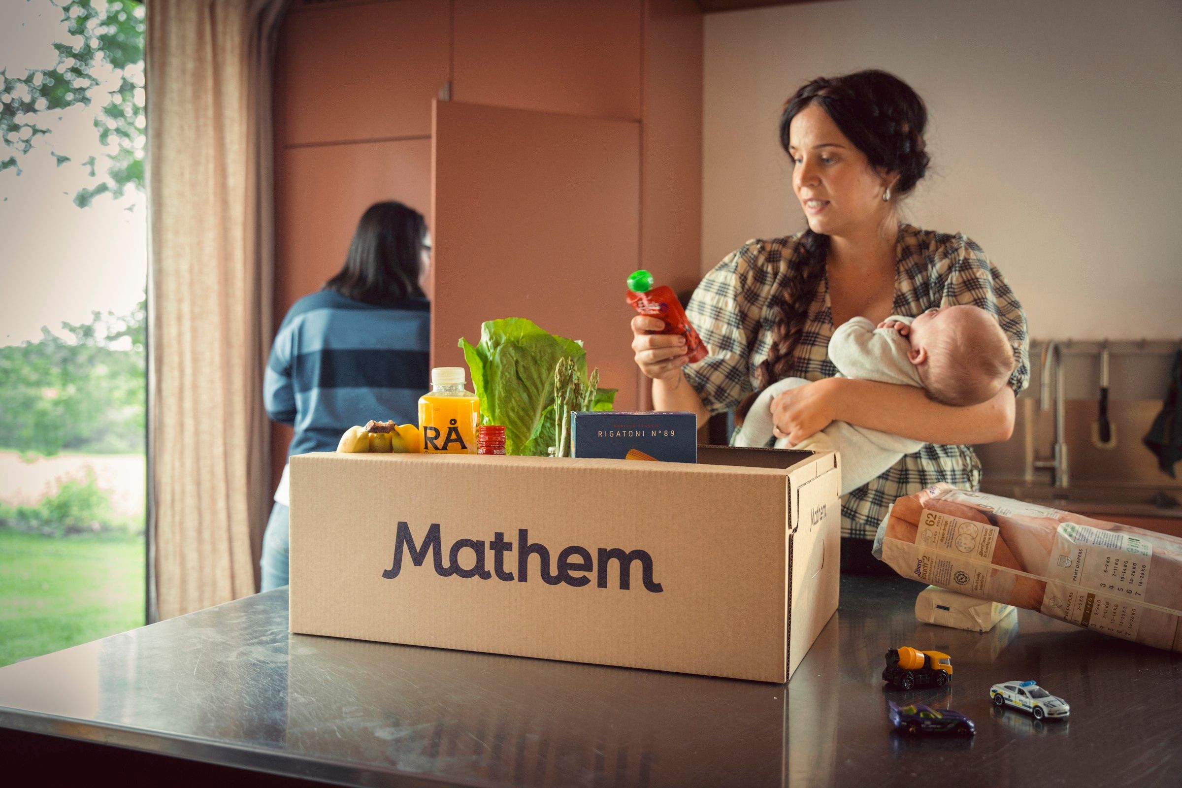 Kvinna håller i bebis och plockar upp en klämmatspåse ur Mathemlåda