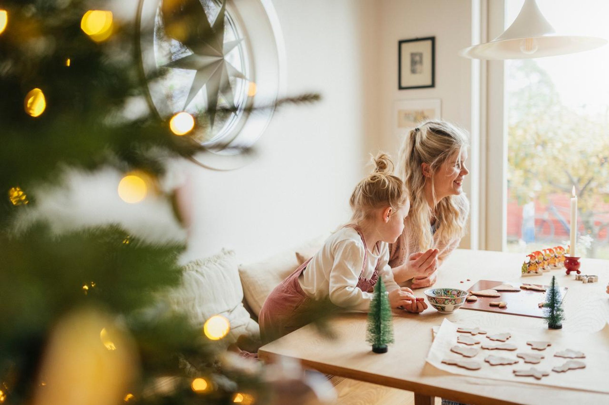 På bildet ser man et barn og en voksen som sitter ved et kjøkkenbord og pynter pepperkaker. I bakgrunnen ser man et pyntet juletre.