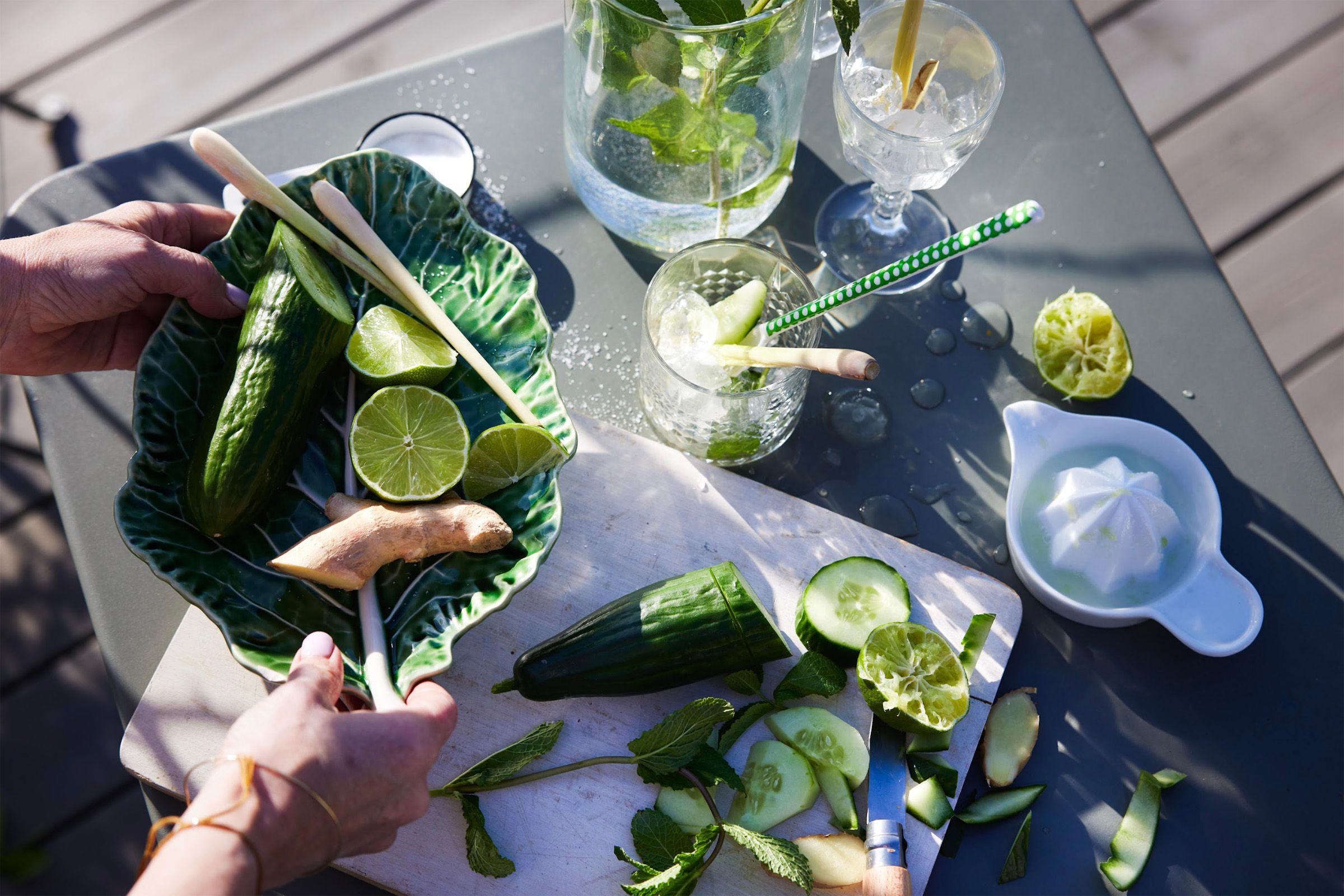 På bildet er et bord dekket for å lage drinker utendørs. En person står og håndterer agurk og lime.