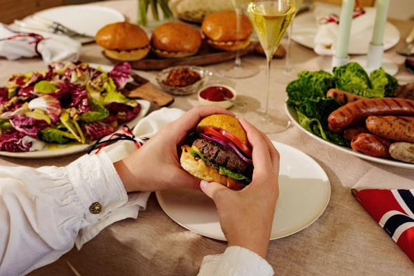 På bildet ser man et matbord dekket til en 17. mai-middag. Det sitter en person ved bordet og holder i en hamburger.
