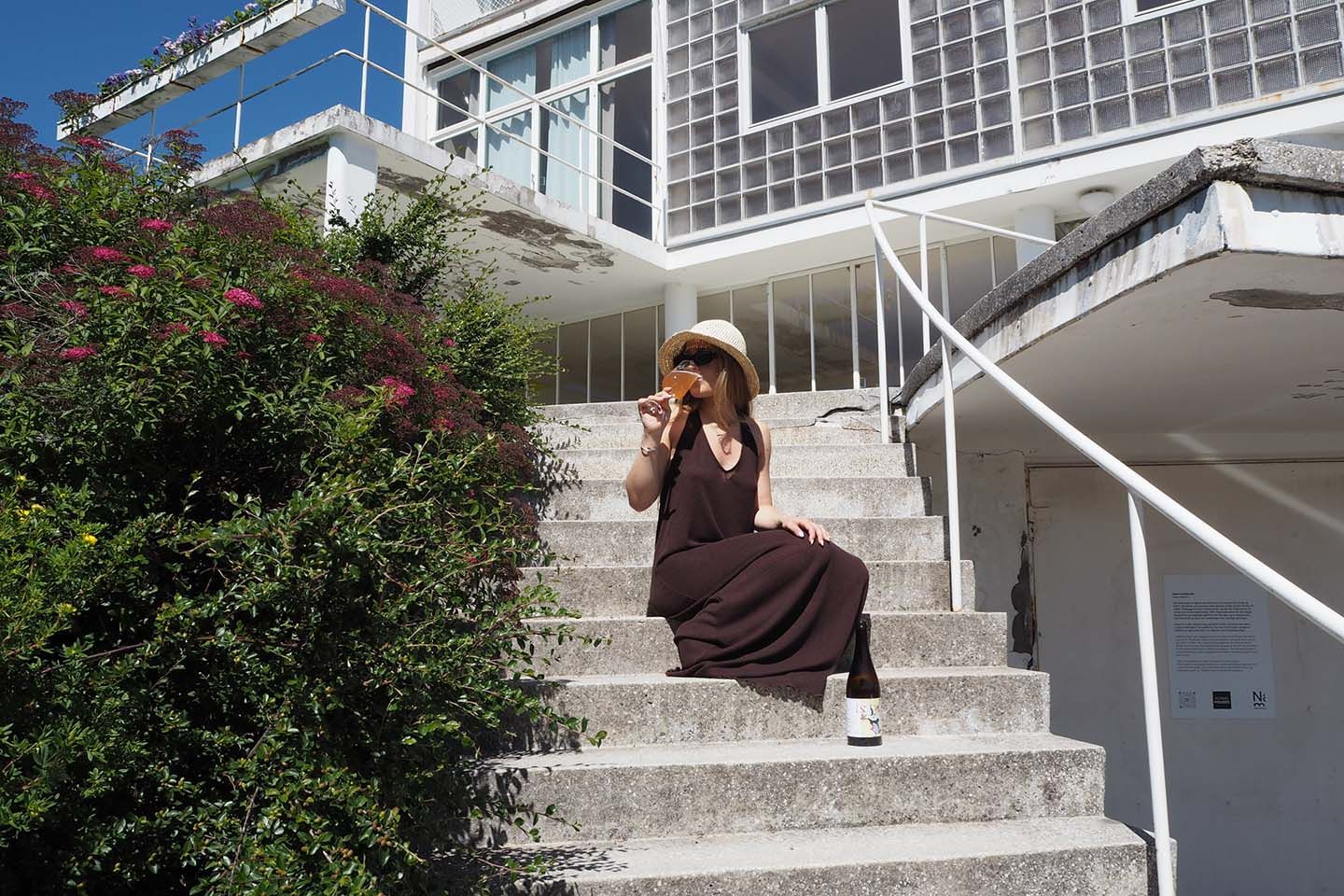 På bildet ser man en kvinne i brun kjole sitte på en betongtrapp utendørs og drikke av et glass, det er sommer.