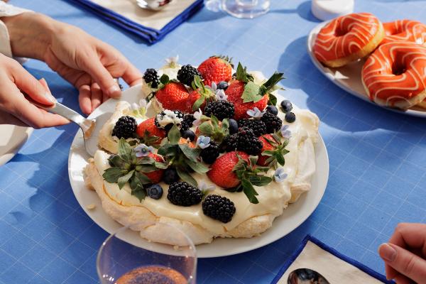 På bildet ser man en pavlova med ferske bjørnebær og jordbær som står på en blå duk. En person gjør seg klar til å ta et stykke av kaken.