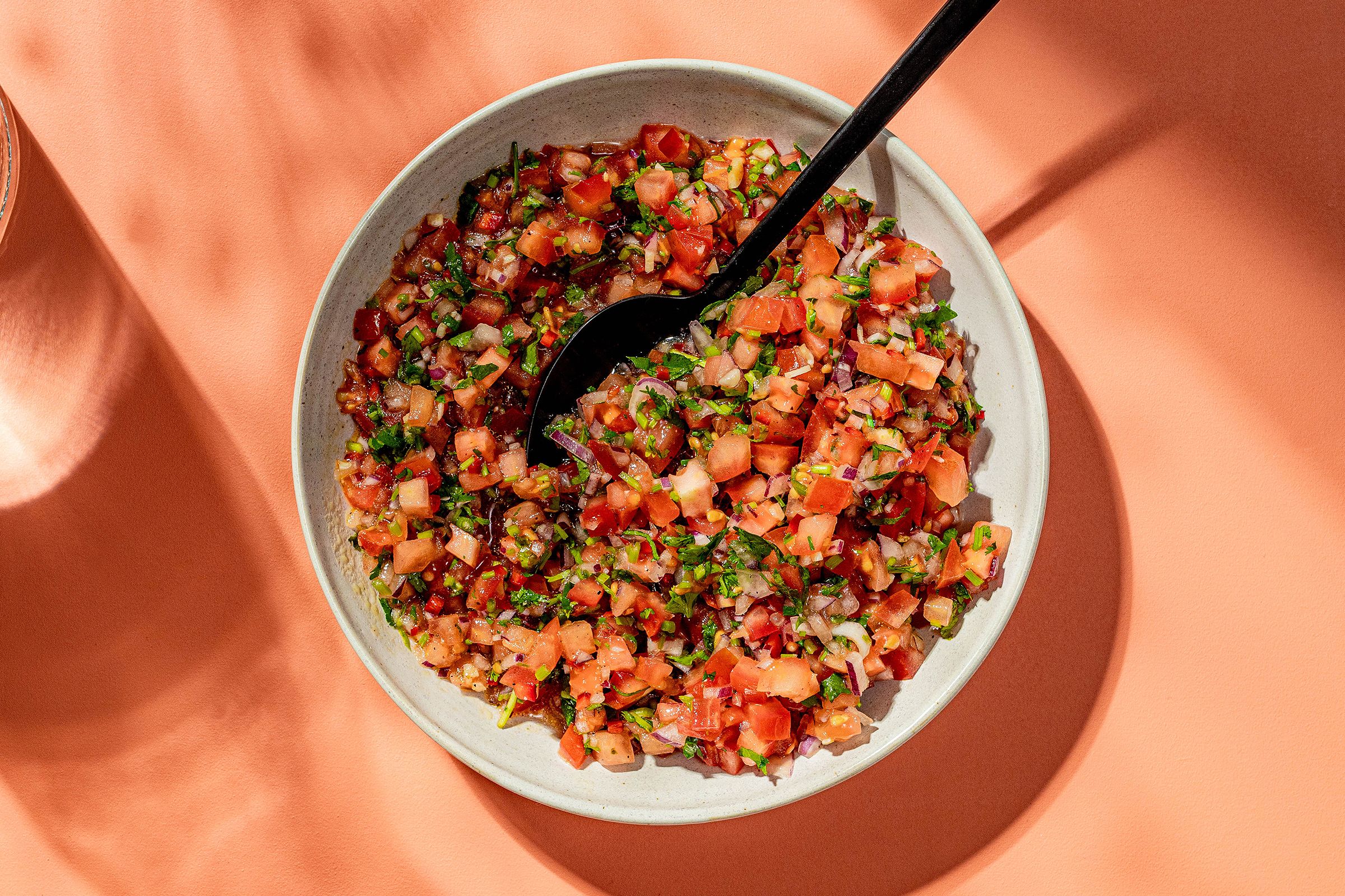 En bolle med pico de gallo står på et ferskenfarget underlag, solen skinner inn fra siden.