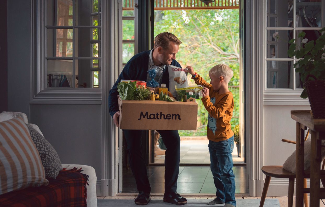 En vuxen person står framför en ytterdörr och håller i en Mathem-låda fylld med varor. Ett barn står bredvid och undersöker innehållet i lådan.