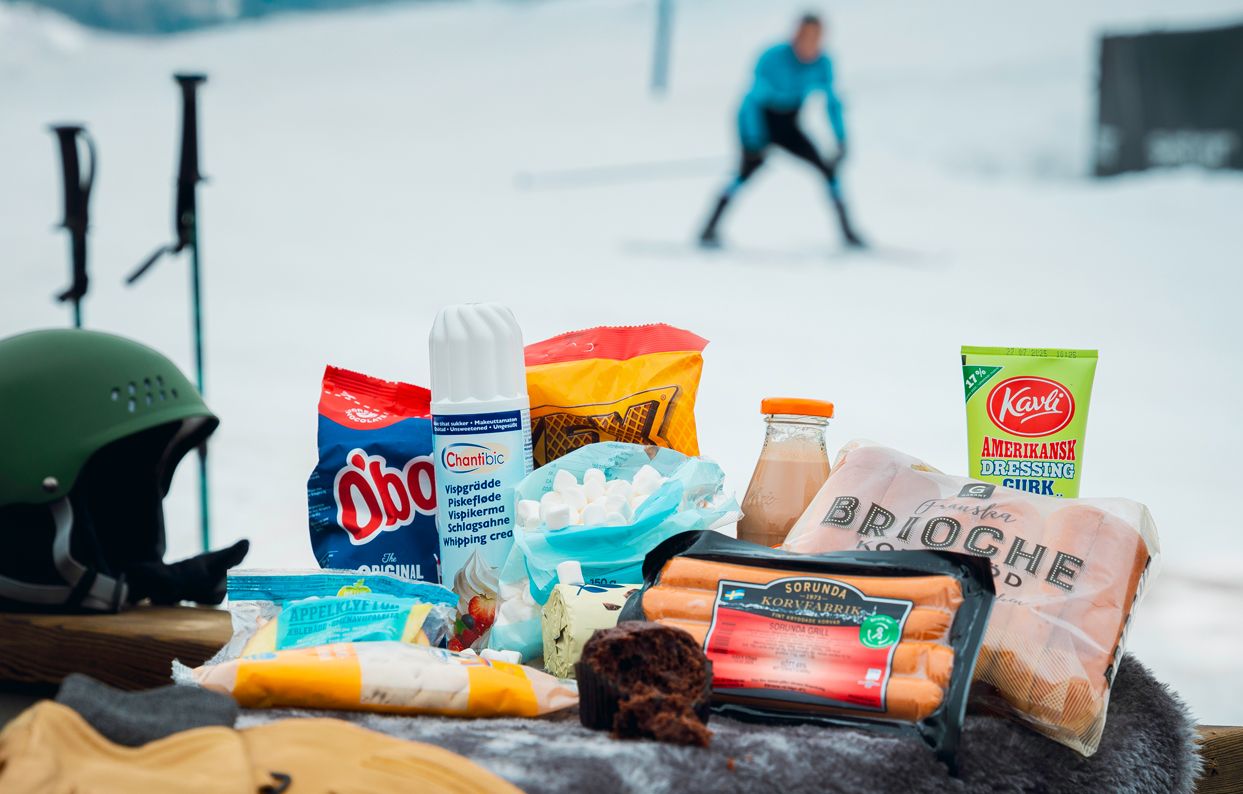 Utvalda produkter upplagda framför en skidbacke. I bakgrunden åker en person skidor nedför backen. Känsla av uteliv och sportlov.