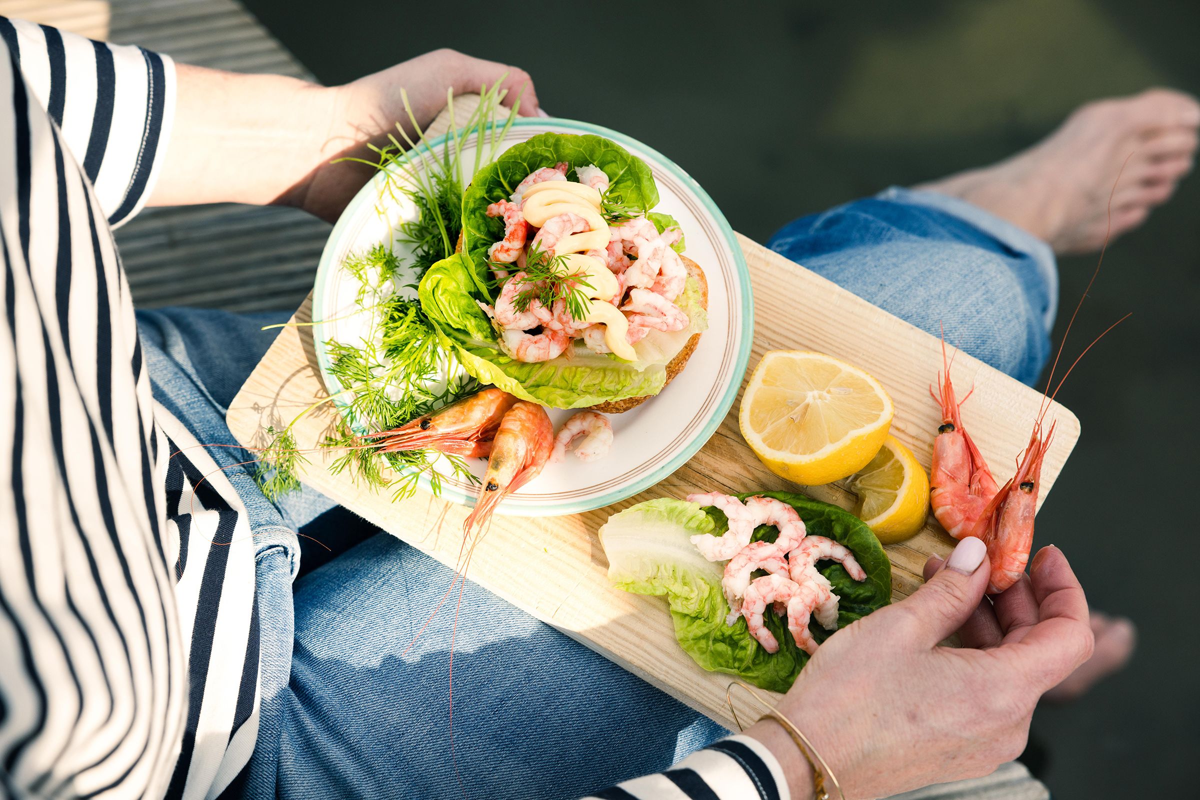 På bildet ser man en person som sitter utendørs på en brygge med et fat på fanget. På fatet har personen et rekesmørbrød.