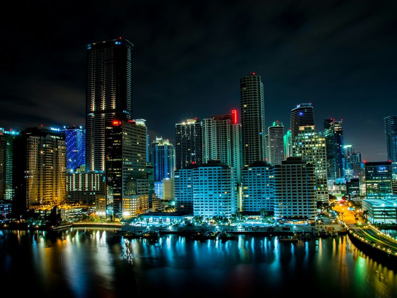 Real photograph of the Miami skyline illuminated at night, reflected across Biscayne Bay