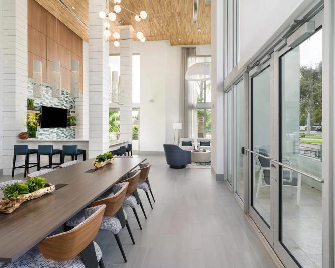 Double height resort clubhouse lounge with a wood plank ceiling, blue tile coffee bar, long communal table with wood chairs, a seating nook under a dome pendant, and full height glass doors opening to a covered patio.