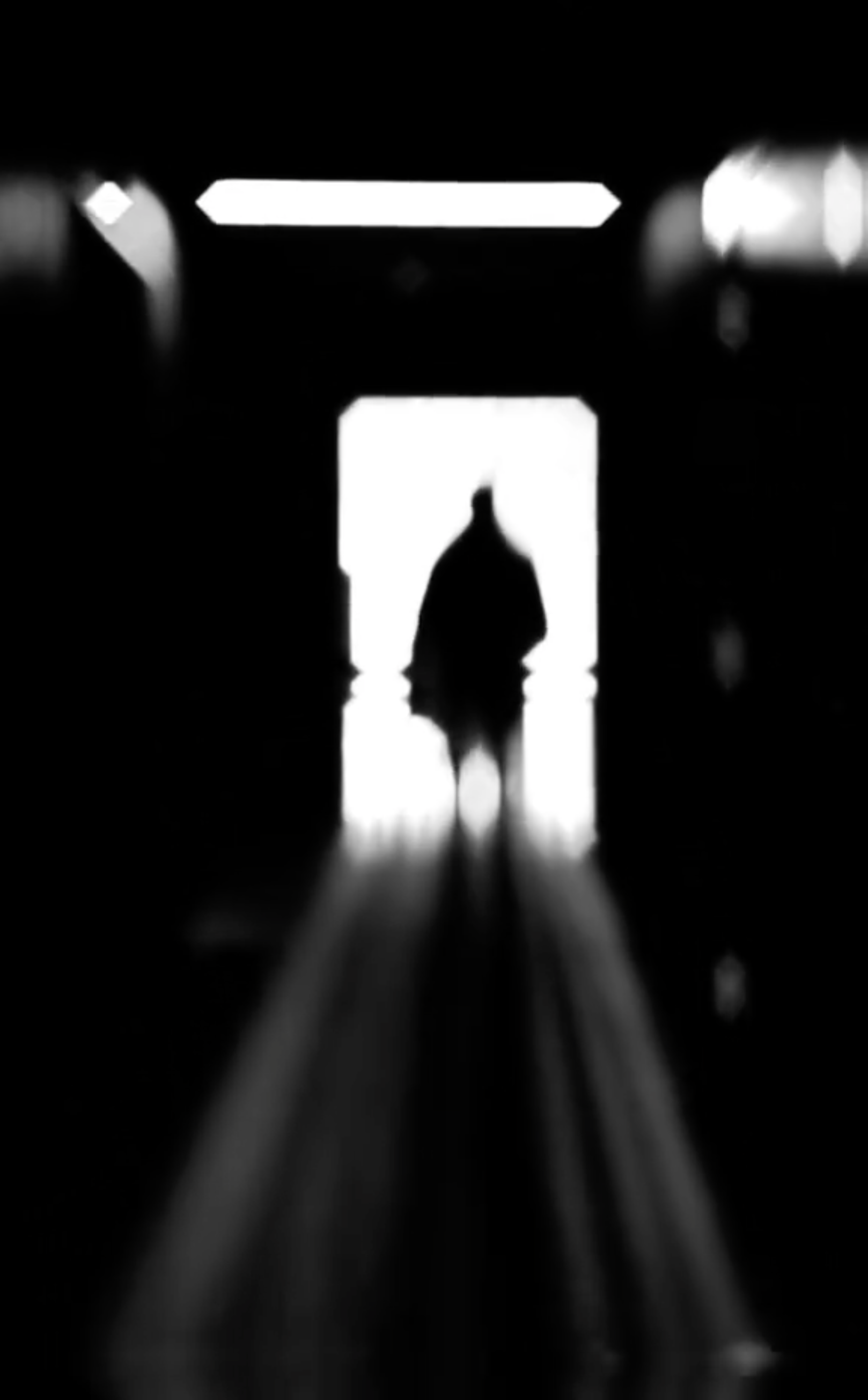 Monochrome moving-image artwork showing a silhouetted human figure travelling through a dark architectural space toward a luminous rectangular opening, surrounded by deep shadows and stark contrasts of light and darkness.