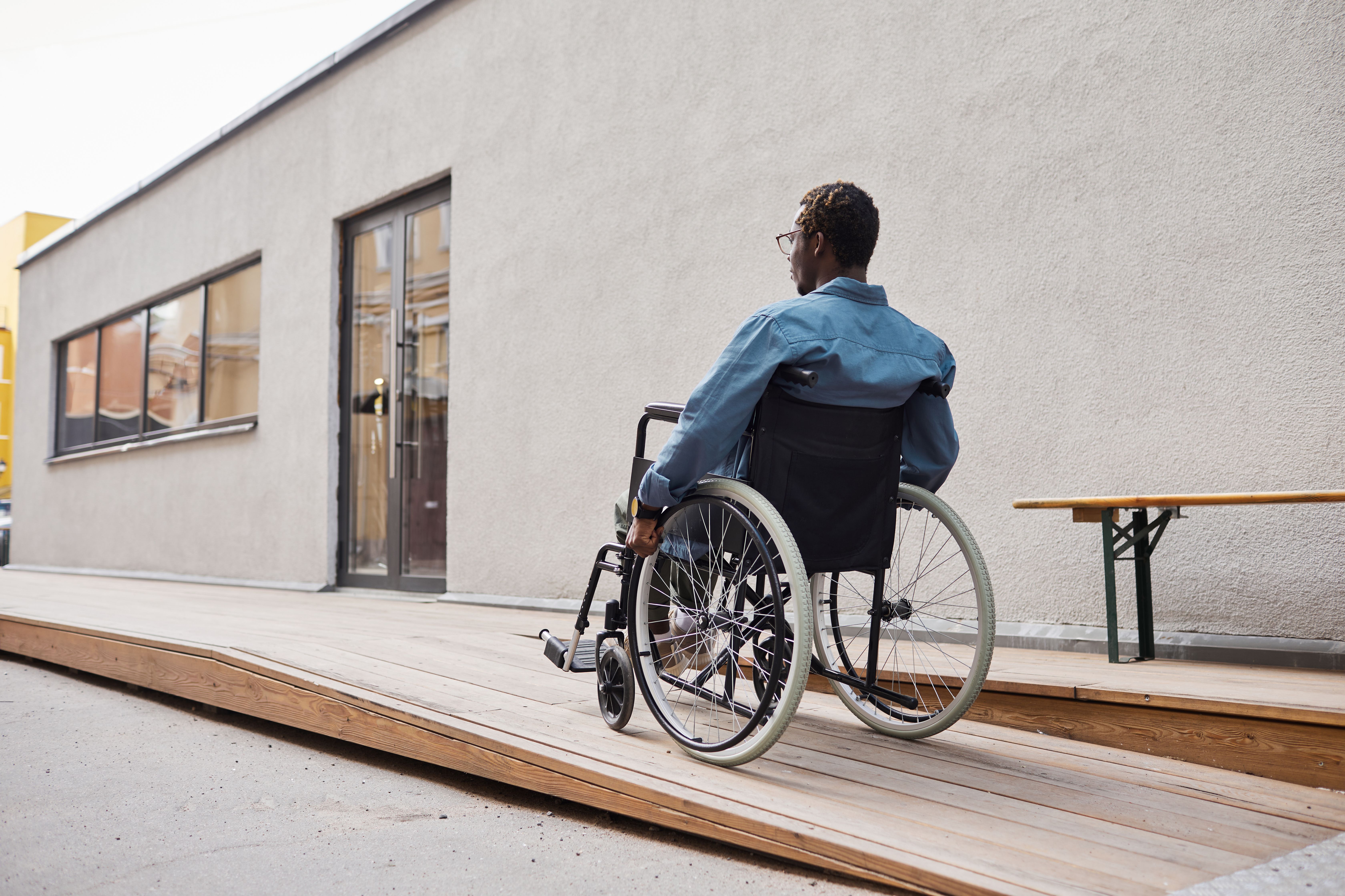 Man in wheelchair going up ramp towards front door of a building
