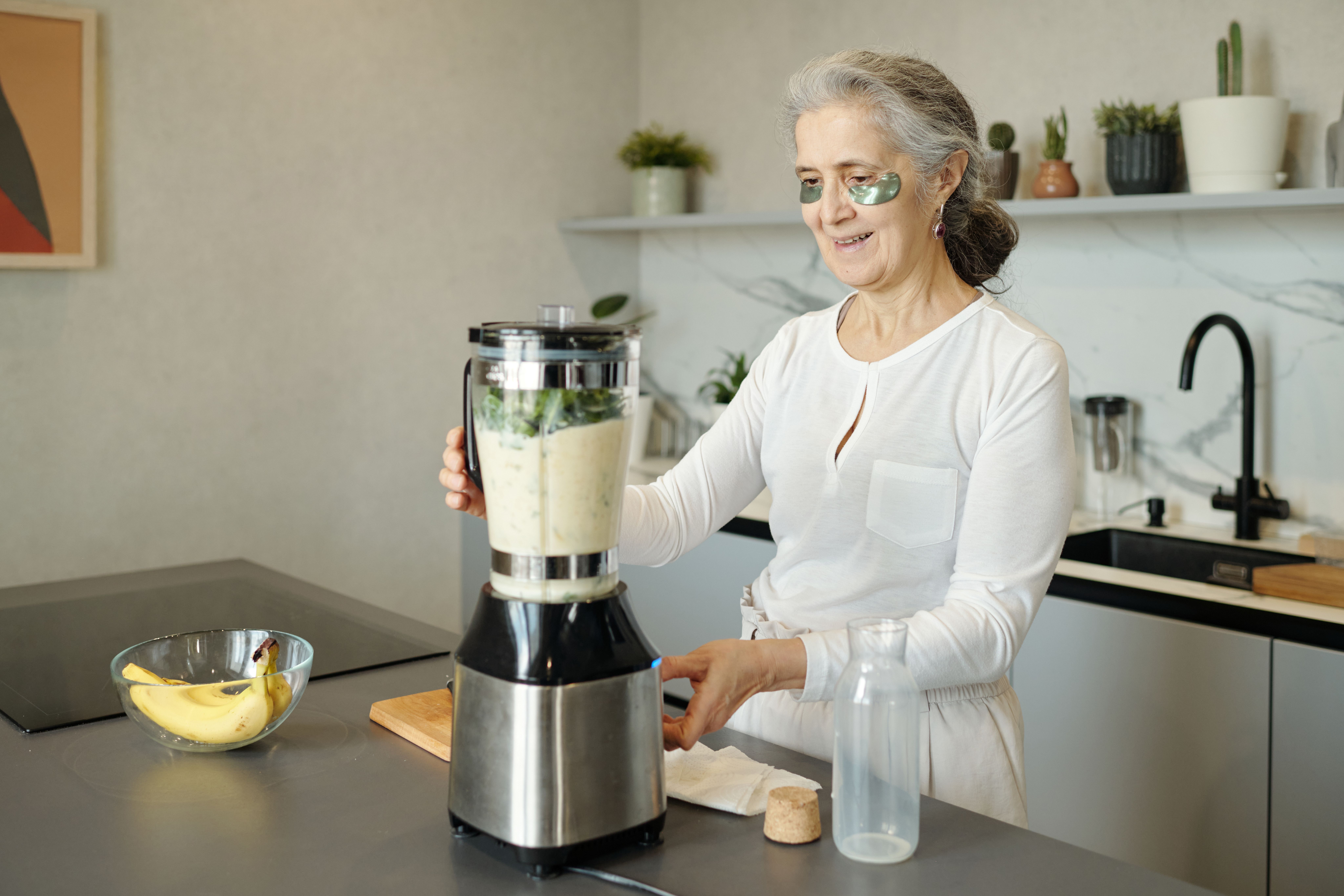 old lady standing in kitchen in front of blender making a banana smoothie