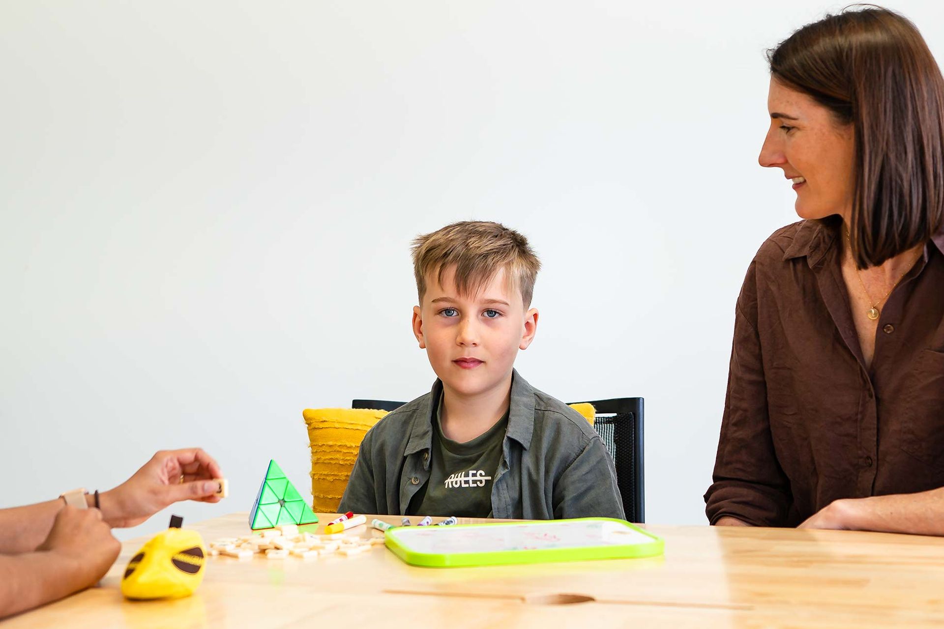 Smiling boy sitting at a table next to his mother with toys and cards in front of him