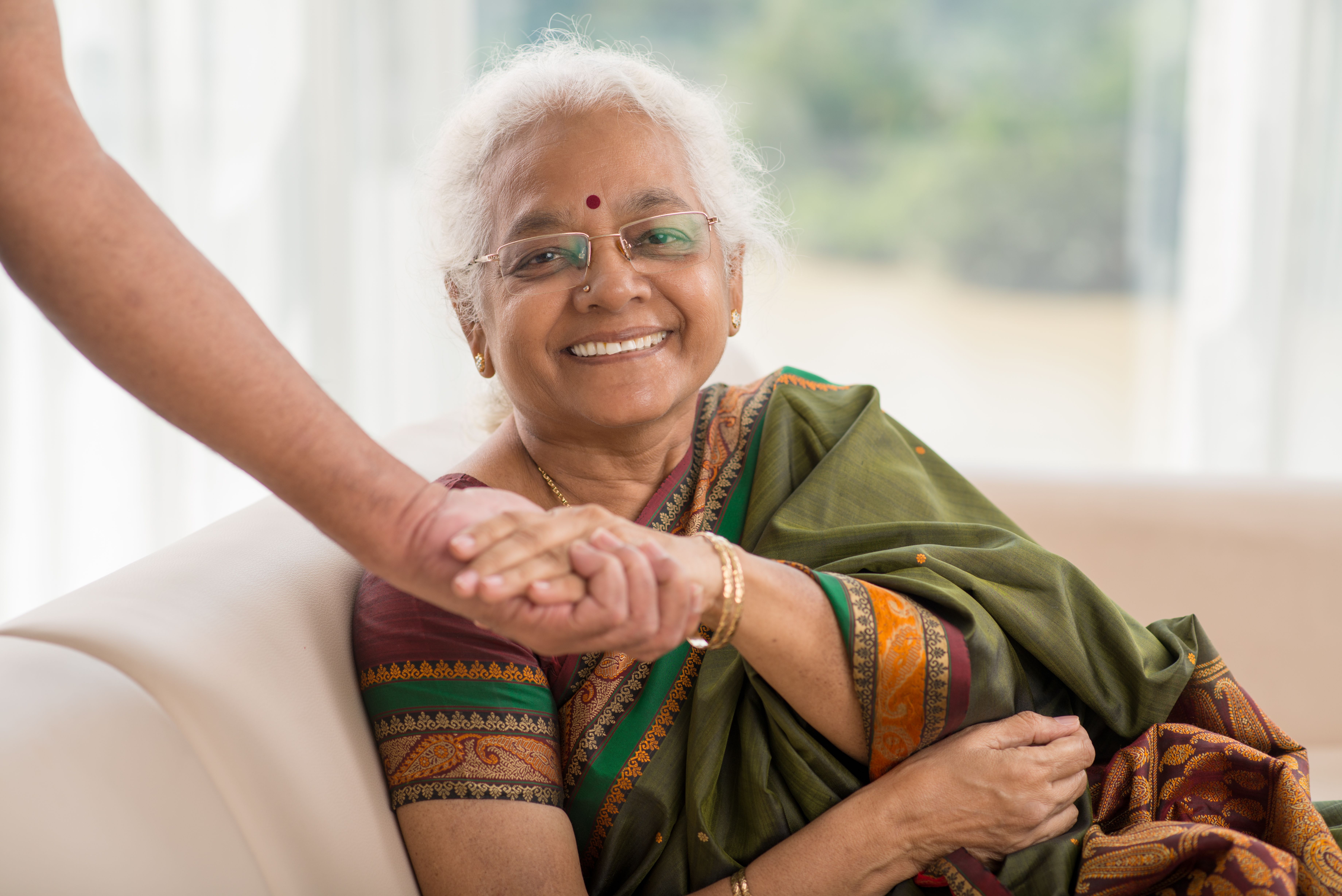 Smiling elderly Indian woman wearing traditional sari sitting on sofa and holding someone's hand.