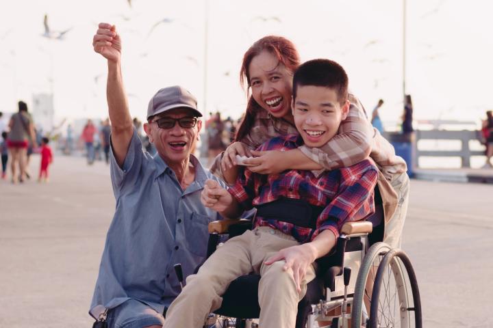 young smiling boy in wheelchair being hugged from behind by smiling woman with smiling man crouching alongside