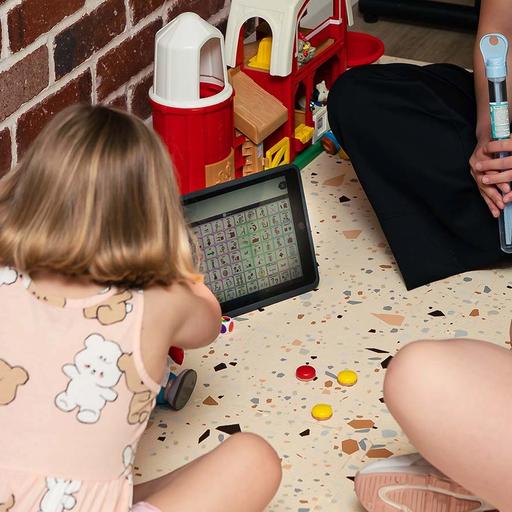 A young girl looking at a tablet, sitting on the floor with another older girl and a BlueRocket clinician