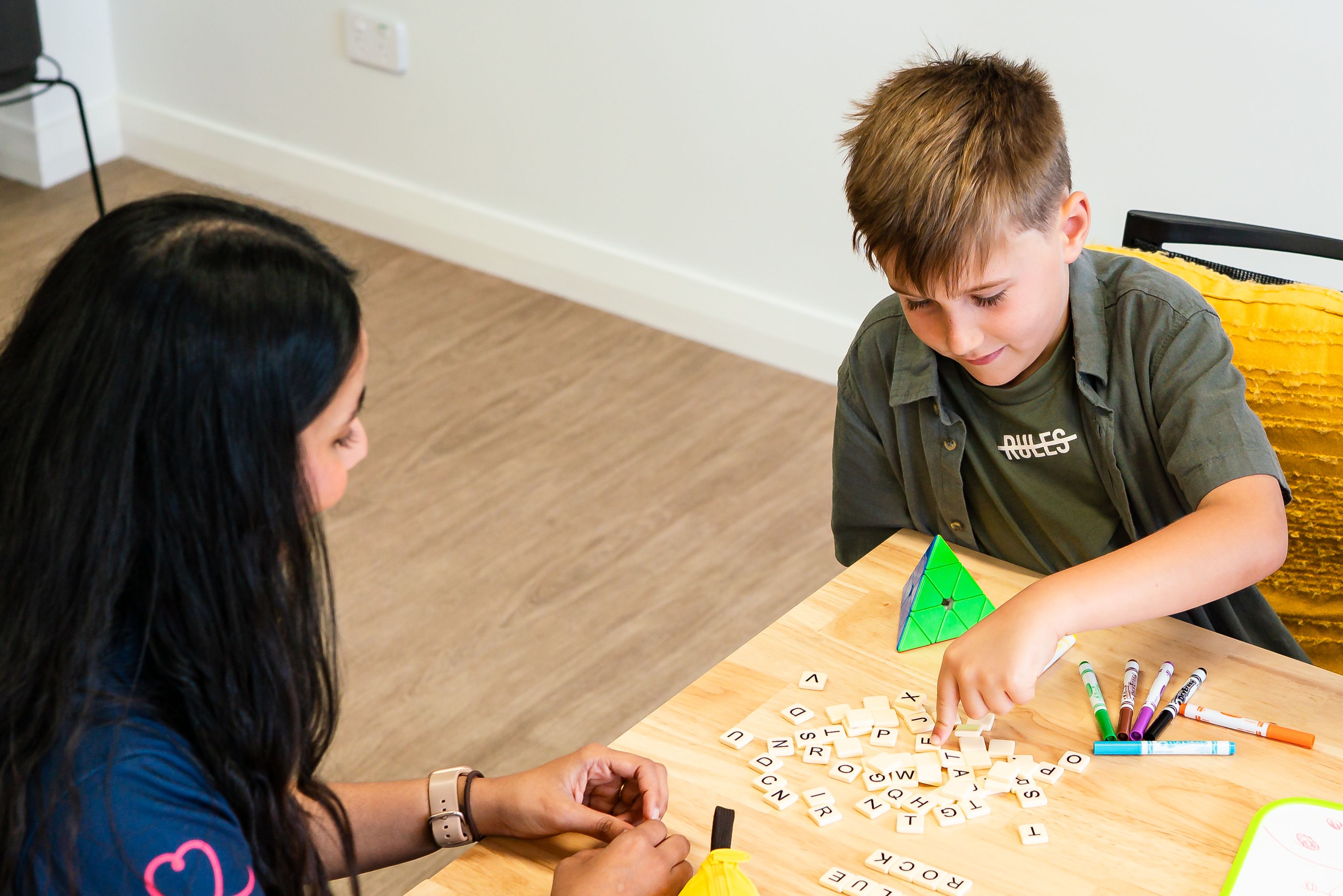A child and a clinician sitting at a table both smiling. The child is spelling the word BlueRocket using scrabble tiles.