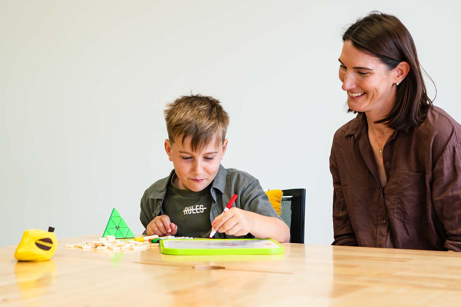 a young boy drawing and playing with letter tiles at a desk beside his smiling mother